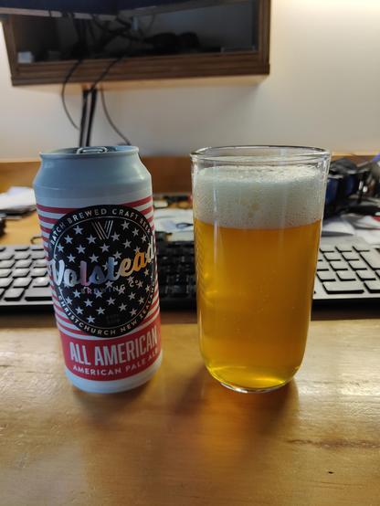 A 440ml beer can with US flag colours & stars and stripes, with the brewery name "Volstead" above the 'All American' beer name and 'American Pale Ale' designation standing next to a similarly tall glass filled with delicious light amber beverage, both on my computer desk. A faint entrancing hint of floral hops is in the air... 